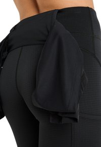 Zwarte sportlegging met een gestructureerd ontwerp met ribbels en een gathered, lichtgewicht overlay bij de taille voor extra bedekking.