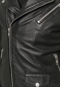 Giacca in pelle nera con cerniere argento sulla parte anteriore principale e sulle tasche, più un dettaglio con patta a bottone a pressione.