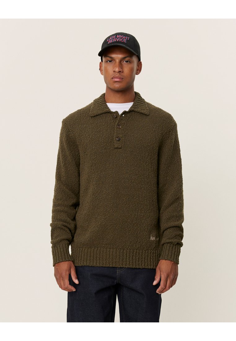 Olivgrön stickad tröja med texturerad yta, krage, treknapprad och ribbade ärmar. Bärs med mörka jeans och en svart keps.