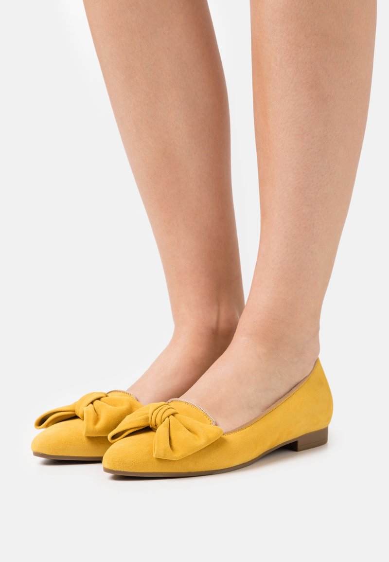 Chaussures plates en daim jaune avec grands nœuds décoratifs portées pieds nus sur un fond blanc.