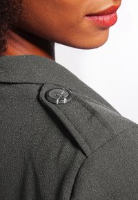Gros plan sur l'épaule d'une personne portant un manteau texturé foncé avec une épaulette boutonnée et ses lèvres visibles de profil.