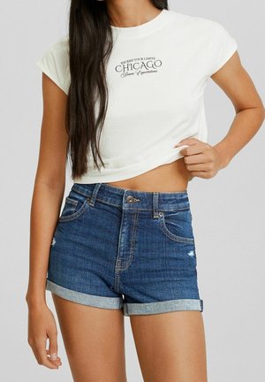 Kvinde iført hvid kort crop-top med teksten "Chicago" og blå denimshorts med opkørt kant, der holder skjortens kant let løftet mod grå baggrund.