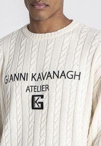 Cremefarbener Kabelstrickpullover mit geripptem Muster und schwarzem gesticktem Logo auf der Vorderseite. Klassischer Rundhalsausschnitt mit lockerer Passform.