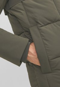 Giacca imbottita verde oliva con tasca con zip e polsini a coste. Presenta una superficie liscia e opaca e una trama trapuntata.