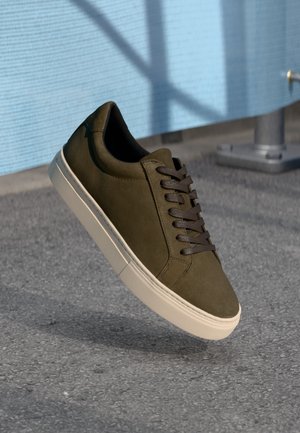 Mørk olivengrønn semsket sneakers med brune snørebånd, kremfarget gummisåle, rund tå og glatt tekstur mot en grå fortausbakgrunn.