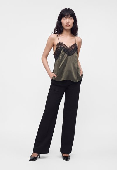 Femme debout, les mains dans les poches, portant un caraco en satin vert olive avec dentelle noire et un pantalon large noir avec des talons noirs.