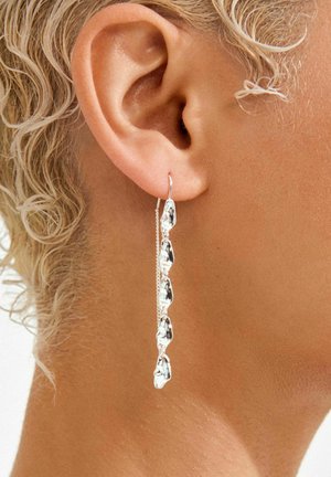 Srebrna viseča uhan z podolgovatimi, kapljičastimi obeski in nežno verižico. Nošen na levem ušesu na teksturirani koži.