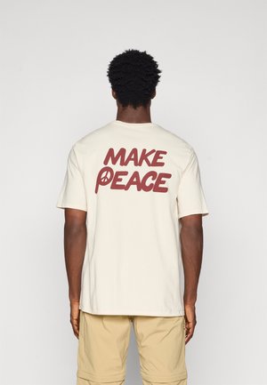 Kortslevar kremfarget t-skjorte med stor brun tekst "MAKE PEACE" på baksiden, med et fredssymbol integrert i bokstaven "P".