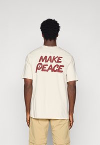 Šortsvārki krēmkrāsas T-krekls ar lielu brūnu tekstu "MAKE PEACE" uz aizmugures, kurā miera simbols ir integrēts burtā "P".