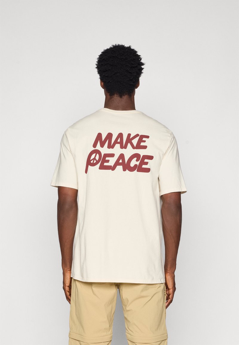 Šortsvārki krēmkrāsas T-krekls ar lielu brūnu tekstu "MAKE PEACE" uz aizmugures, kurā miera simbols ir integrēts burtā "P".