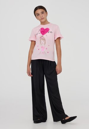 T-shirt rosa con maniche a volant, caratterizzato da una grafica a cuore e un'illustrazione di ballerina. I pantaloni neri, ampi e fluidi, completano l'outfit.