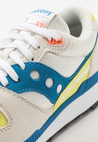 Ljusgrå sneakers med blåa och gula detaljer. Har en överdel i mocka, mesh-tyg och vita snören. Logotyp på tungan. Strukturerad sula.