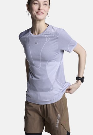 Vrouw die een lichtpaarse sportshirt verstelt, draagt een bruine korte broek en een zwart fitnesshorloge, glimlacht en kijkt opzij.