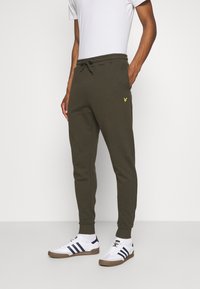 Mann trägt olivgrüne Jogginghosen mit gelbem Logo auf dem Oberschenkel, weiße Socken, weiße Sneakers mit navyblauen Streifen und ein weißes T-Shirt.