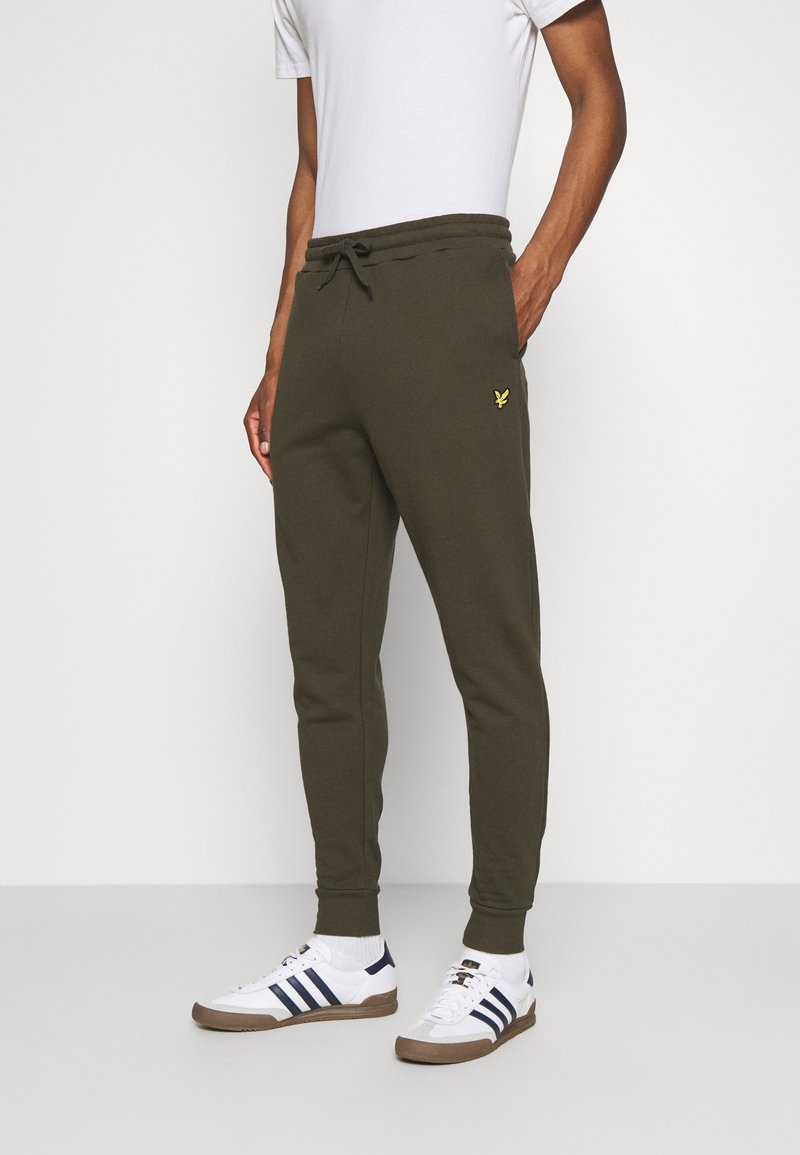 Mann trägt olivgrüne Jogginghosen mit gelbem Logo auf dem Oberschenkel, weiße Socken, weiße Sneakers mit navyblauen Streifen und ein weißes T-Shirt.