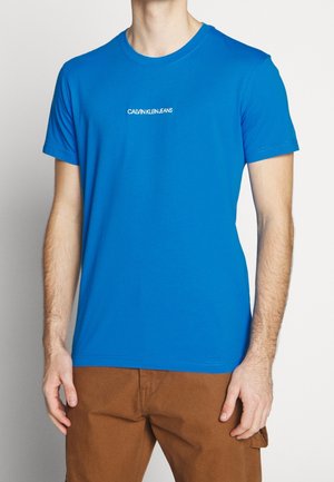 T-shirt en coton bleu à manches courtes, col rond, avec le logo blanc "CALVIN KLEIN JEANS" centré sur la poitrine. Texture lisse et design simple.