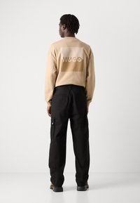 Beige sweatshirt with a large HUGO logo in tan on the back; paired with black cargo pants featuring side pockets and a relaxed fit.