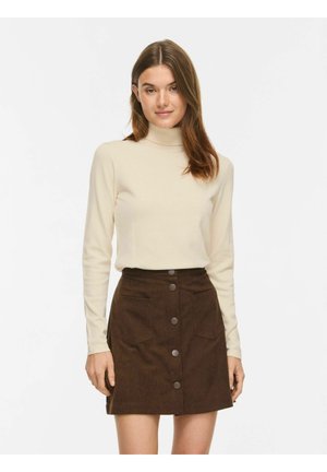 Femme portant un pull à col roulé crème et une jupe marron à boutons sur le devant avec poches, debout devant un fond uni.
