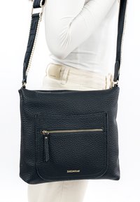 Person trägt ein weißes Oberteil und helle Hosen und hat eine strukturierte schwarze Leder-Umhängetasche mit Frontreißverschlusstasche und verstellbarem Riemen bei sich.