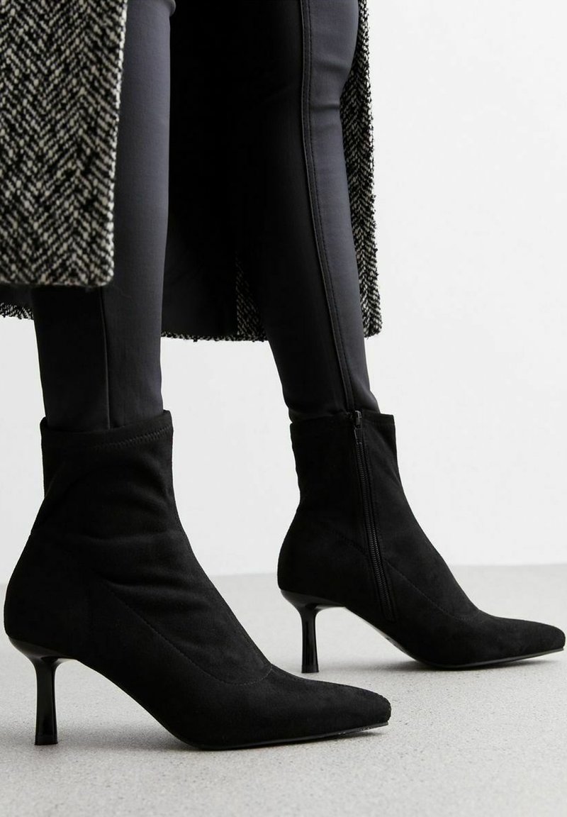 Bottines en daim noir à bout pointu avec petits talons, portées avec un pantalon slim noir et un manteau à motif noir et blanc.