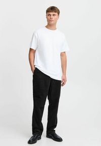 Camiseta blanca de algodón con un corte relajado, combinada con pantalones de pana negros. El modelo lleva zapatos de cuero negros, con las manos en los bolsillos.