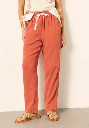 Žena oblečená v voľných hrdzavej farby textúrovaných nohaviciach s bielou šnúrkou a viacfarebných sandáloch, stojaca pri neutrálnej stene.