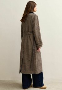 Abrigo de trench de cuadros marrón y negro, ceñido en la cintura, con un panel lateral marrón oscuro en contraste. Llevado sobre jeans de pierna ancha y zapatos beiges.