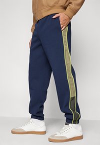 Calças de fato azul-marinho com uma faixa lateral em verde-oliva e amarelo neón. Modelagem tipo jogger com punho no tornozelo. Combinadas com ténis brancos.