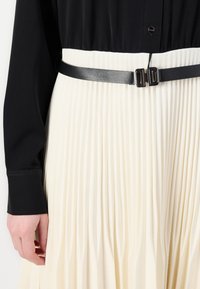 Blouse noire à manches longues associée à une jupe crème plissée, cintrée à la taille par une fine ceinture noire avec une double boucle.