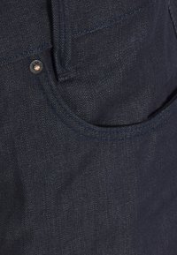 Vaqueros de mezclilla oscura con un bolsillo delantero, costuras reforzadas y un botón metálico decorativo. La tela tiene una textura y brillo sutiles.