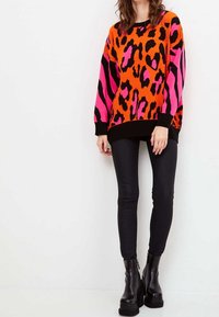 Kolorowy, oversize'owy sweter w pomarańczowym, różowym i czarnym kolorze z wzorami lamparta i zebry, zestawiony z czarnymi legginsami i masywnymi czarnymi butami.