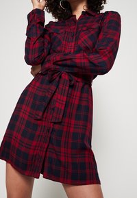 Rode en marineblauwe geruite jurk van flanel, met lange mouwen, twee borstzakken, knoopsluiting aan de voorkant en een ceintuur rond de taille.