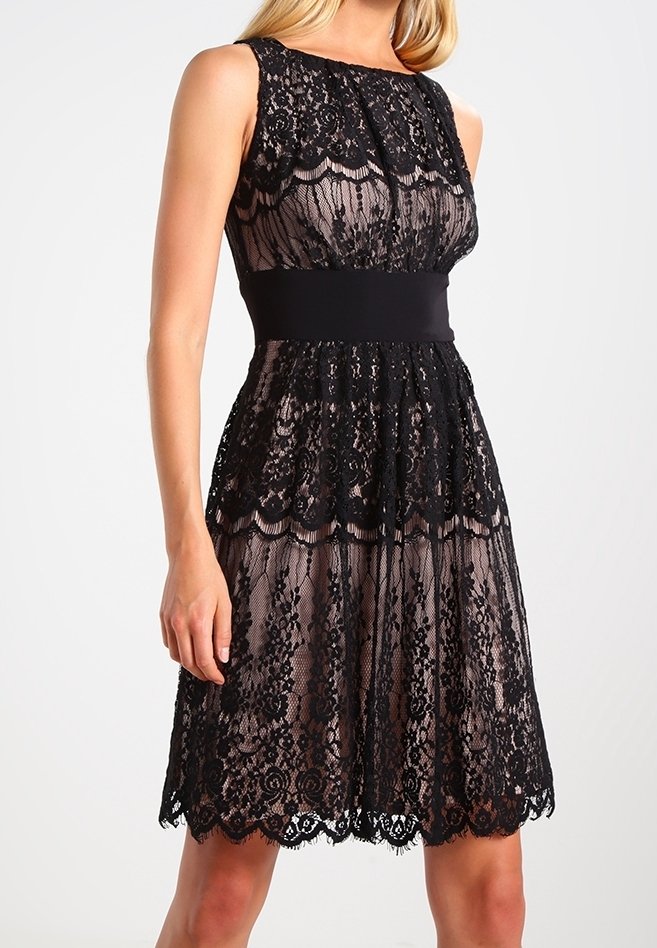 Robe en dentelle noire avec un buste ajusté, un ourlet en forme de vagues et une ceinture noire lisse. Le tissu semi-transparent révèle des motifs floraux élaborés.