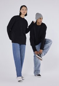 Două modele poartă tricouri lungi supradimensionate negre; unul în blugi denim de culoare albastră deschisă, celălalt cu o căciulă gri și sneakers negri. Fundal neutru.