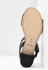 Sandale noire avec dessus en cuir lisse, bride ajustable et semelle plate. Présente une semelle extérieure beige avec une surface texturée antidérapante et un détail de logo.