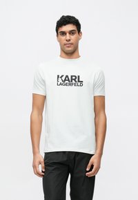 Λευκό κοντομάνικο μπλουζάκι με μαύρο λογότυπο "KARL LAGERFELD". Λείο βαμβακερό ύφασμα με στρογγυλή λαιμόκοψη, φορεμένο με μαύρο παντελόνι.
