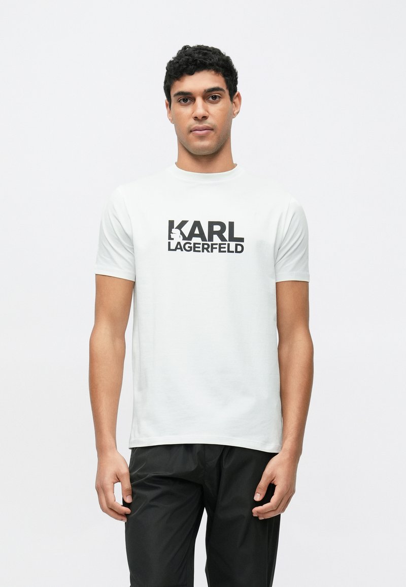 Λευκό κοντομάνικο μπλουζάκι με μαύρο λογότυπο "KARL LAGERFELD". Λείο βαμβακερό ύφασμα με στρογγυλή λαιμόκοψη, φορεμένο με μαύρο παντελόνι.