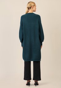 Cardigan a coste color turchese con fitting rilassato, maniche lunghe e tasche laterali, abbinato a pantaloni larghi neri con pois bianchi e tacchi.