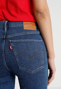 Tmavě modré džíny z denimu s hnědou koženou záplatou na zadní straně, výrazným dvojitým šitím na kapsách a červenou štítkovou etiketu na boku.