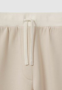 Beige Jogginghose mit einem gerippten, elastischen Bund, einer Kordelzugvorrichtung und einer Seitentasche. Der Stoff hat eine weiche, glatte Textur.
