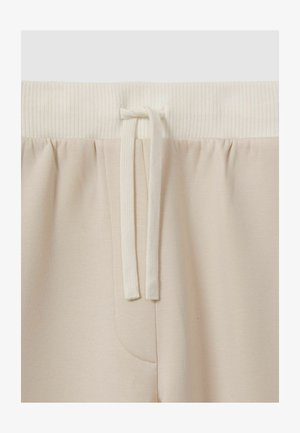 Pantaloni della tuta beige con vita elastica a coste, chiusura con cordino e tasca laterale. Il tessuto ha una texture morbida e liscia.