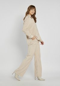 Beżowa koszula z korduroy i pasujące spodnie, o luźnym kroju. Modelka nosi jasne czółenka na wysokim obcasie; jednolite tło.