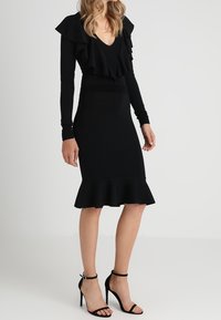 Robe noire à col volanté, manches longues et silhouette cintrée. Elle présente un ourlet évasé et des chaussures à talons hauts avec bride à la cheville.