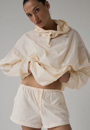 Jasno-kremowy, lekki sweter z kapturem, elastycznymi mankietami i sznureczkiem. Zestawiony z pasującymi szortami z elastycznym pasem.