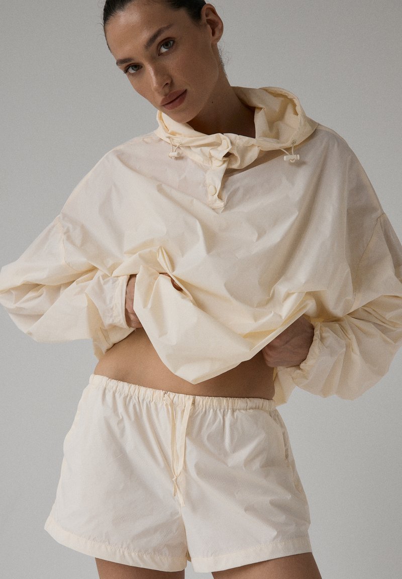 Jasno-kremowy, lekki sweter z kapturem, elastycznymi mankietami i sznureczkiem. Zestawiony z pasującymi szortami z elastycznym pasem.