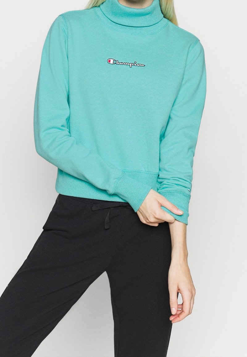 Felpa turtleneck turchese con polsini e orlo a costine. Presenta un logo ricamato in rosso e bianco. Abbinata a pantaloni neri.