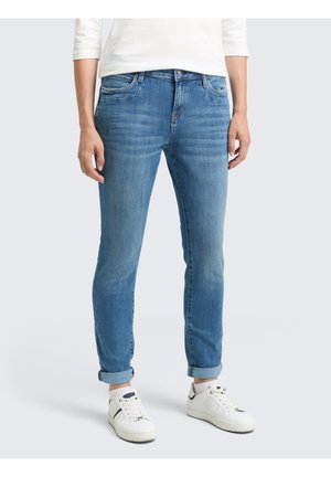 Personne portant un jean bleu slim à revers et des baskets blanches, debout, les mains détendues le long du corps, sur un fond uni.