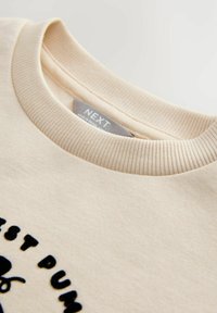 Cremfarbener Sweatshirt mit einem gerippten Halsausschnitt, versehen mit schwarzem gesticktem Text. Glatte Textur mit einem lässigen Design. Etikett innen sichtbar.