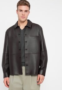 Barna bőrkabát, galléros nyakkal, gombos záródással és zsebekkel. Sima textúrájú, laza fazonú, sötét pulóver felett viselve.