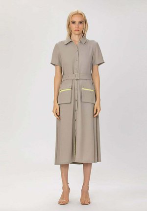 Femme portant une robe midi beige boutonnée, manches courtes, taille ceinturée, deux grandes poches devant et des talons ouverts beiges.